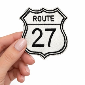 May include: A shield-shaped embroidered patch featuring a white background with a black border. The text ROUTE is stitched in black at the top, and the number 27 is prominently displayed in large black lettering in the center of the patch.
