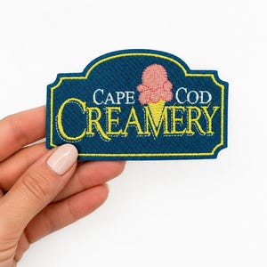May include: A rectangular embroidered patch with a dark blue background and a yellow border. The text reads CAPE COD CREAMERY in white and yellow lettering. A pink ice cream scoop on a yellow cone is positioned above the word CREAMERY.