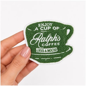 May include: A green embroidered patch shaped like a coffee cup with white stitched text that reads ENJOY A CUP OF Ralph's COFFEE JAVA & MOCHA. The patch features a textured surface and a white border, designed for application to fabric items.