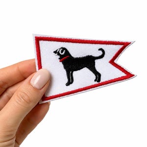 May include: A triangular pennant-shaped embroidered patch featuring a white background with a red border. In the center, there is a black dog silhouette with a red collar. The patch has a clean, stitched edge and a flat, fabric-like texture.