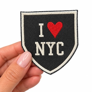 May include: A shield-shaped embroidered patch with a black background and a white border. The patch features the white text I, a red heart symbol, and the white text NYC stacked below. The design is stitched with a textured fabric finish.