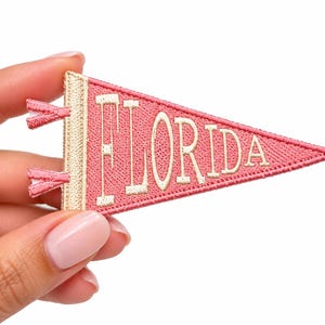 May include: A triangular pennant-shaped embroidered patch in pink with a cream-colored border and two small ties on the left side. The word FLORIDA is stitched in cream-colored thread across the center of the pink fabric surface.