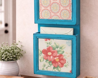 Op de afbeelding: Een turquoise wandpostorganizer met twee compartimenten. Het bovenste gedeelte heeft een patroon, terwijl de onderste een bloemenprint toont. Een witte envelop ligt in het onderste compartiment. De organizer is van hout.