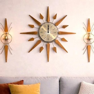 Puede incluir: Un salón con un sofá gris, cojines coloridos y una mesa de centro de madera. Un reloj de estrella y apliques de pared a juego adornan la pared. Una lámpara y una estantería completan la escena, creando un ambiente acogedor.