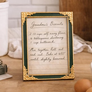 May include: A framed recipe for Grandma's Biscuits with a gold and green frame. The recipe includes ingredients like self-rising flour and shortening. The image also shows a bowl of flour, eggs, and a white pitcher, suggesting a baking theme.