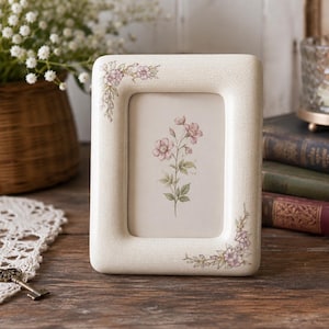 Puede incluir: Un marco de fotos rectangular de color crema con un sutil acabado craquelado. El marco presenta delicadas ilustraciones florales rosas en las esquinas superior izquierda e inferior derecha. Un estampado botánico a juego se muestra dentro.