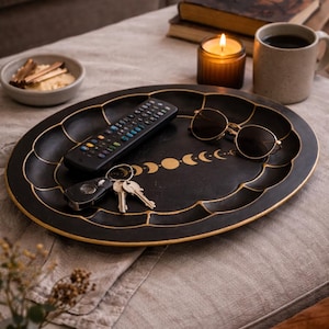 May include: Black oval tray with gold trim and a crescent moon design. The tray holds a remote control, keys, and sunglasses. A lit candle, coffee mug, and a small bowl are also visible.
