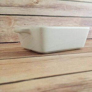 May include: A small, rectangular white ceramic baking dish with a handle on one side. The dish is set on a light brown wooden surface. The dish is empty and appears clean.