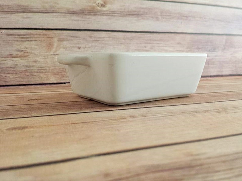 May include: A small, rectangular white ceramic baking dish with a handle on one side. The dish is set on a light brown wooden surface. The dish is empty and appears clean.