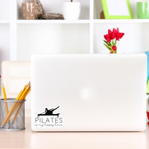 May include: A white laptop computer with a black and white sticker that says "PILATES is my happy hour". The sticker features a silhouette of a person doing a pilates exercise. The laptop is on a light wood table with a vase of pink flowers, a pencil holder with yellow pencils, and a small potted cactus.