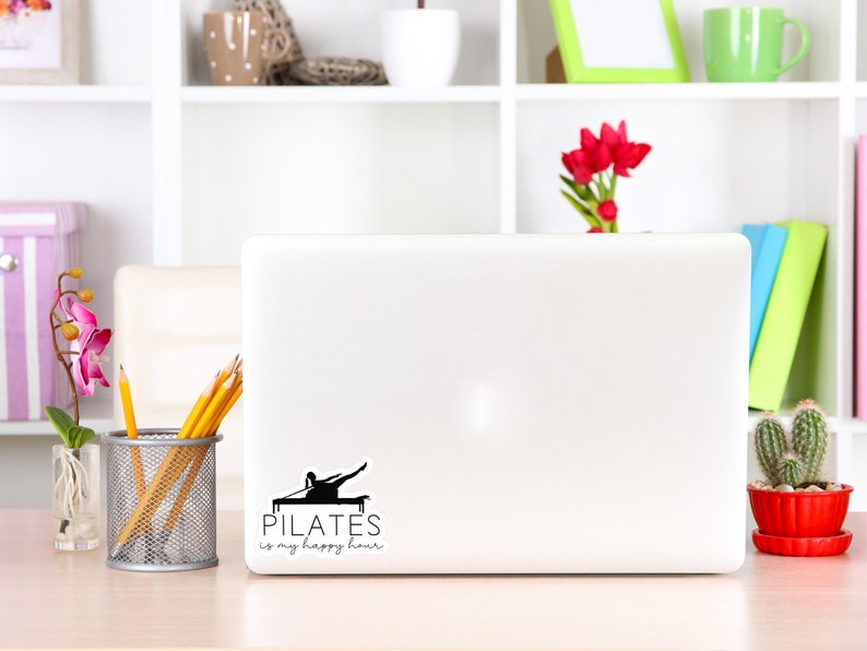May include: A white laptop computer with a black and white sticker that says "PILATES is my happy hour". The sticker features a silhouette of a person doing a pilates exercise. The laptop is on a light wood table with a vase of pink flowers, a pencil holder with yellow pencils, and a small potted cactus.