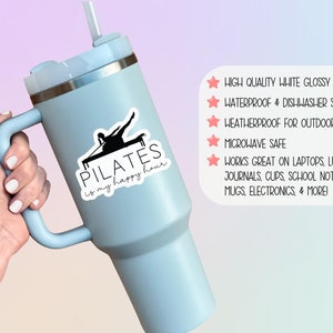 May include: A blue stainless steel tumbler with a black and white vinyl sticker that says "PILATES is my happy hour". The sticker features a silhouette of a person doing pilates on a reformer. The tumbler has a handle and a straw.