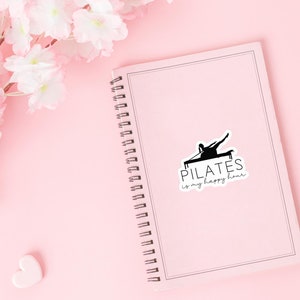 May include: A pink notebook with a spiral binding and a black and white sticker that says "PILATES is my happy hour". The sticker features a silhouette of a person doing pilates on a reformer.