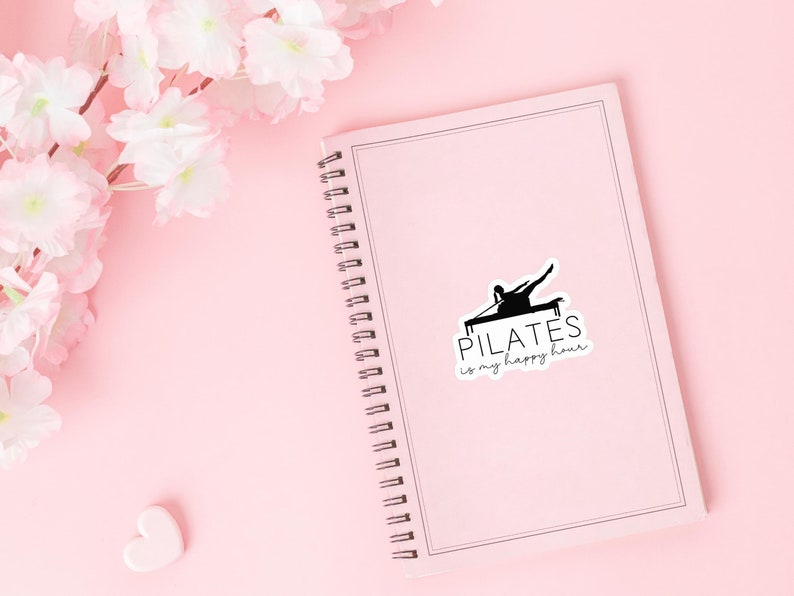 May include: A pink notebook with a spiral binding and a black and white sticker that says "PILATES is my happy hour". The sticker features a silhouette of a person doing pilates on a reformer.