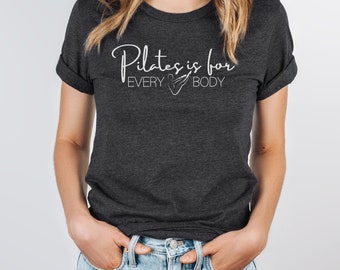 Peut inclure: Un t-shirt gris foncé avec du texte blanc qui dit "Pilates is for EVERY BODY" avec une image stylisée d'une personne faisant du pilates.