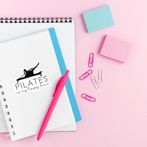 May include: A white notebook with a black and white sticker that says "PILATES is my happy hour" and a black silhouette of a person doing pilates. The notebook is on a pink surface with a pink pen, blue notebook, and pink and blue sticky notes.