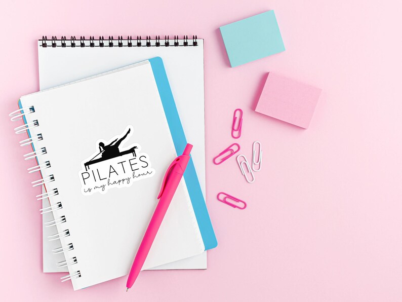May include: A white notebook with a black and white sticker that says "PILATES is my happy hour" and a black silhouette of a person doing pilates. The notebook is on a pink surface with a pink pen, blue notebook, and pink and blue sticky notes.