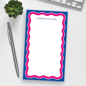 May include: A personalized notepad with a blue border and a wavy pink inner border. The text "audrey reynolds" is printed at the top. A silver pen and a small succulent plant are next to the notepad.