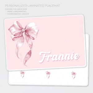 May include: A personalized laminated placemat with a pink bow design and the name "Frannie" in white script. The placemat has a pink diamond pattern background and the text "PERSONALIZED LAMINATED PLACEMAT" at the top.