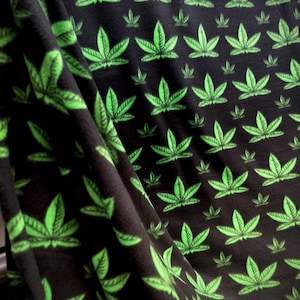 May include: Black fabric with a repeating pattern of bright green cannabis leaves. The leaves are detailed with veins and are arranged in a grid-like fashion. The fabric is draped, showing folds and shadows.