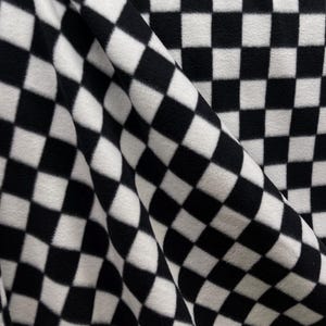 May include: Close-up of a black and white checkered fabric. The fabric is soft and appears to be fleece or a similar material. The pattern is a classic checkerboard design, with each square being a uniform size. The fabric is draped, creating folds and shadows.