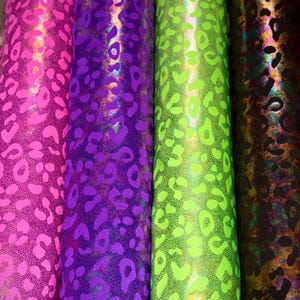 Puede incluir: Cuatro rollos de tela estampada en colores vibrantes. Los rollos muestran un diseño de estampado de leopardo en rosa, morado, verde neón y marrón con reflejos iridiscentes. La tela está enrollada, mostrando el patrón repetitivo.