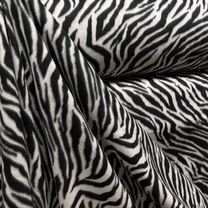 Puede incluir: Primer plano de una tela con un llamativo estampado de rayas de cebra. La tela presenta rayas negras y blancas alternas, creando un diseño de alto contraste. El material parece suave y flexible, con pliegues y drapeados visibles.