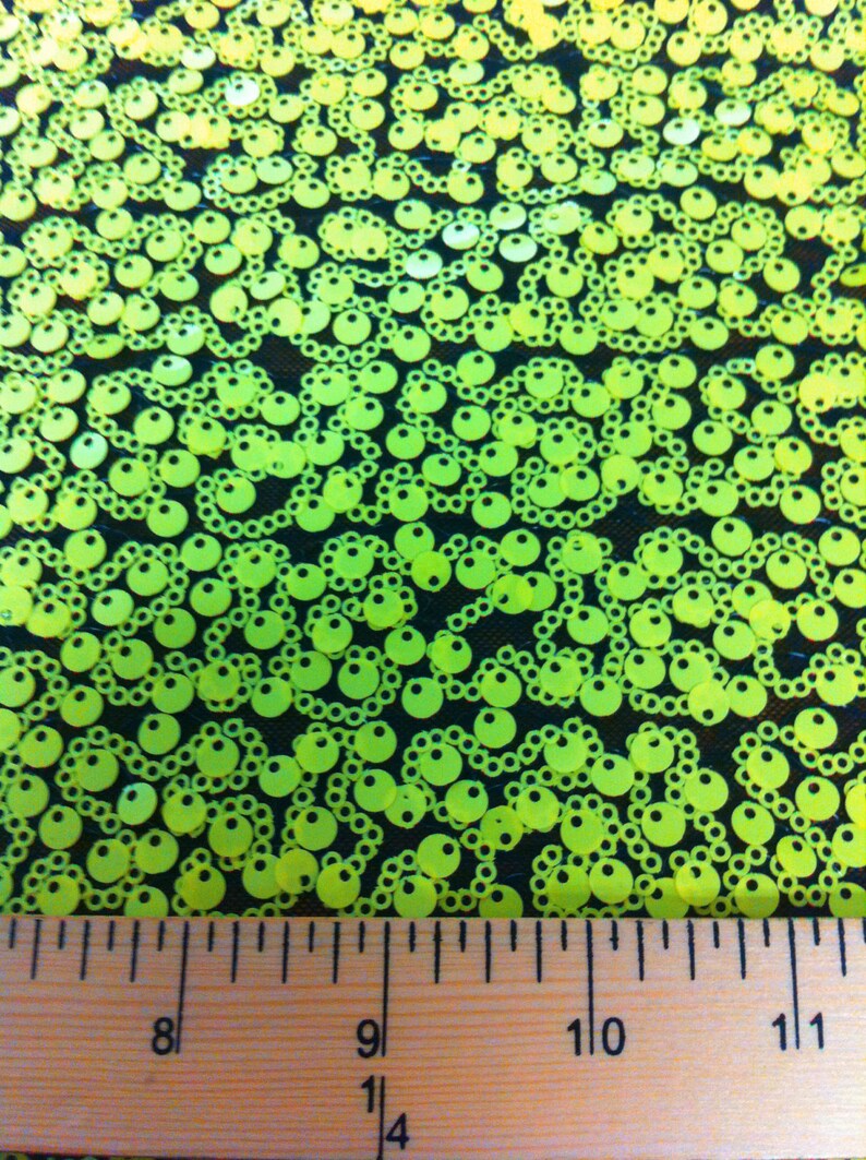 May include: Close-up of neon green sequins on a black mesh fabric. A ruler is placed at the bottom of the image, showing measurements in inches.