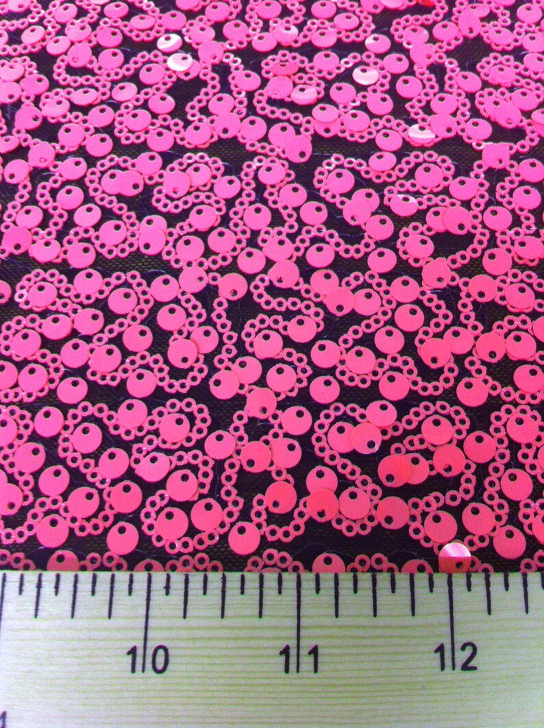 May include: Pink sequins on a black mesh fabric. A ruler is placed at the bottom of the image with the numbers 10, 11, and 12 visible.