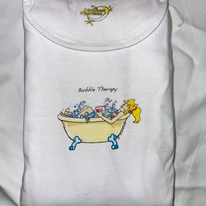 May include: White t-shirt featuring a cartoon illustration of a woman in a bathtub filled with bubbles, holding a glass of wine. The text "Bubble Therapy!" is above the image. The text "Southern Charm Creations" is in the top corner of the shirt.