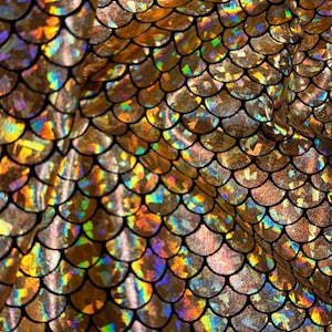 Puede incluir: Una tela con un patrón repetitivo de escamas superpuestas en un acabado dorado metálico. Cada escama individual muestra un brillo holográfico multicolor vibrante que cambia con la luz, delineado por finas líneas curvas de color negro.