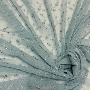 May include: A light blue sheer fabric with a delicate floral pattern embroidered in silver thread. The fabric is draped and gathered, showcasing its texture and drape.