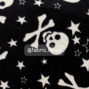 以下が含まれることがあります： 白いドクロと交差した骨と星の模様がついた黒いフリース生地。生地には「@fabric.for.us」というテキストが印刷されています。