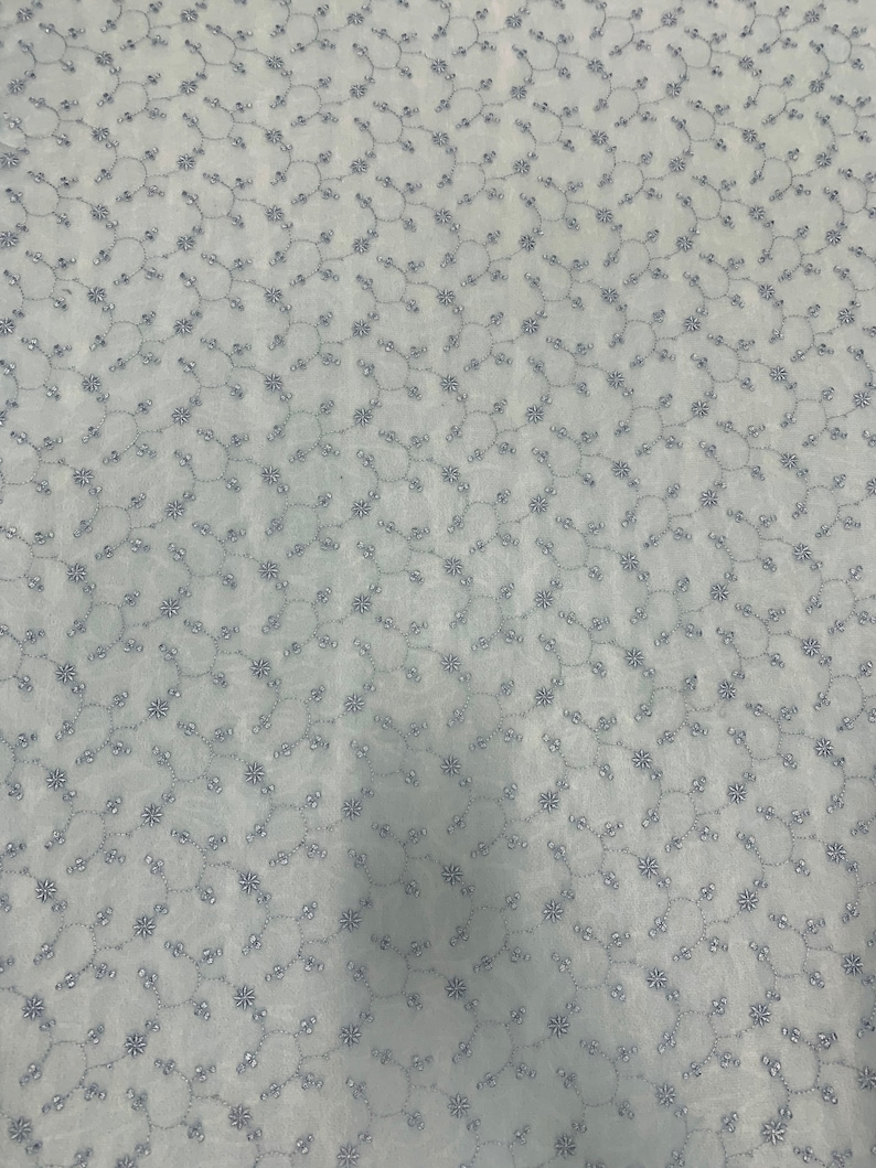 May include: A white fabric with a delicate embroidered pattern of small flowers and vines. The fabric has a subtle sheen and a soft texture.