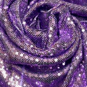 May include: Close-up of a shimmering fabric with a deep purple hue. The material is densely covered in small, round sequins that catch the light, creating a sparkling effect. The fabric is draped and folded, showing its texture and flow.