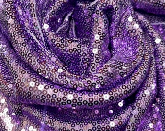 May include: Close-up of a shimmering fabric with a deep purple hue. The material is densely covered in small, round sequins that catch the light, creating a sparkling effect. The fabric is draped and folded, showing its texture and flow.