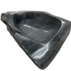 May include: A black marble dish with a smooth, rounded interior and a rough, textured exterior. The dish is shaped like a shallow bowl and has a slightly irregular shape.
