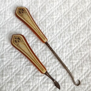May include: Two antique metal tools with brown and white handles. The tools have intricate designs on the handles and are used for sewing or crafting.