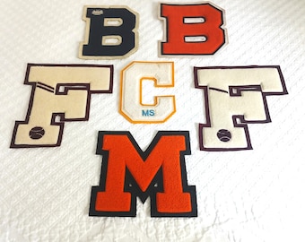 May include: A collection of six varsity-style felt patches featuring letters and symbols. The set includes a navy B, an orange B, two cream F patches with dark brown trim and baseball graphics, a cream C with orange trim and MS text, and an orange M.
