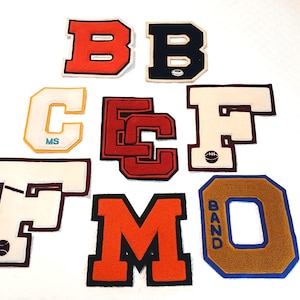 May include: A collection of eight varsity-style felt letter patches in various colors including orange, black, white, red, and gold. The letters include B, B, C, E, F, F, M, and O, with some featuring small embroidered icons like a football or baseball.
