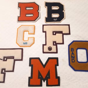 May include: A collection of six varsity-style felt letter patches in various colors. The patches include the letters B, B, C, F, F, M, and O. Some patches feature small embroidered icons like a football, baseball, or the text MS and BAND.