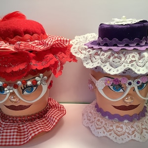 May include: Two decorative flower pots with painted faces, red and white gingham trim, and white lace hats. The red pot has a red and white gingham hat with a red feather. The purple pot has a purple hat with white lace trim.