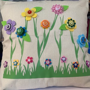May include: A beige pillow with a green grass border and a variety of colorful flowers made from buttons. The flowers are yellow, pink, orange, purple, green, and blue.