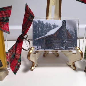 May include: Three small framed pictures on easel stands. The first picture is a cartoon of a girl in a red and black plaid dress. The second picture is a snowy cabin in the woods. The third picture is a green and brown patterned background with a sign that reads "A. Malcolm printer & bookseller".