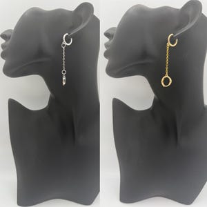 May include: Two earrings displayed on black mannequin heads. One earring is silver with a chain and a small charm. The other earring is gold with a chain and a small circle charm. Both earrings have a hoop that goes through the ear.