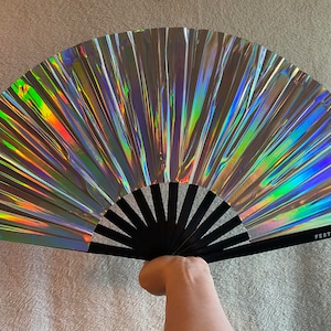 May include: A silver iridescent folding fan with a black handle. The fan has a holographic finish and the handle has the text "FESTV FRILLS" printed on it.