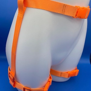 May include: Bright orange harness with adjustable straps and plastic buckles. The harness is displayed on a white mannequin, with straps around the waist and thighs. The background is a solid blue.
