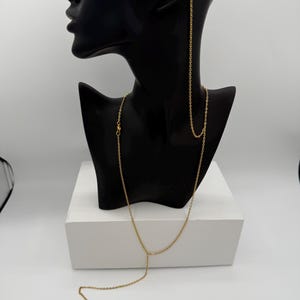 May include: A gold-colored necklace and earring set displayed on a black mannequin head. The necklace features a delicate chain with a Y-shaped design and a double-circle pendant. The earrings are hoop style with a small pearl.