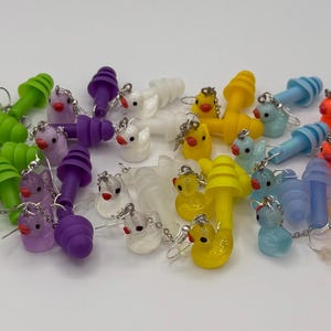 Puede incluir: Una colección de tapones para los oídos de colores con pequeños amuletos de animales. Los tapones para los oídos vienen en varios colores, incluyendo rosa, verde, morado, blanco, amarillo, azul, naranja y turquesa. Cada tapón tiene una pequeña cadena y un amuleto con forma de pato u otro animal.