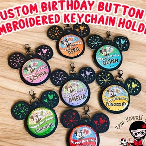 May include: A collection of seven circular birthday buttons attached to black embroidered keychain holders shaped like mouse ears. Each button features a cartoon character, a birthday cake, a name, and an age. The ears are decorated with stitched fireworks and balloon designs in various colours.